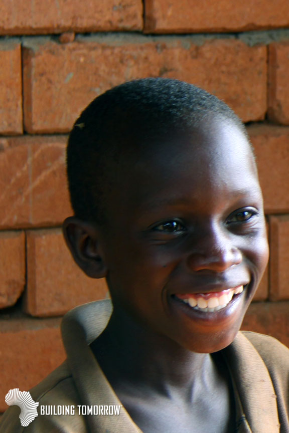 Allan, age 13, is all smiles outside of the first of three classroom blocks for his future school - the Building Tomorrow Academy of Bubeezi, supported by the Engage Network.