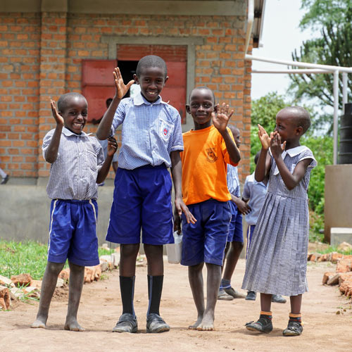Four students wave and laugh on the path outside their school.