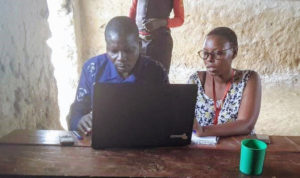 Fellow Namata Tendo sits with second grade (P2) teacher Patrick Byaruhanga at New Eden Primary School as he demonstrates what he has learned. Patrick has never had a chance to use a computer and is excited about being able to touch one for a year through the ICT classes. He hopes the classes will help him to record his information at his farm and provide a creative way for both teachers and students to admire his class. 